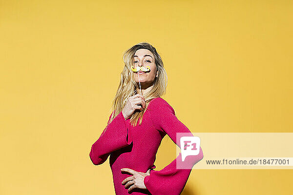 Woman with hand on hip holding fake mustache against yellow background