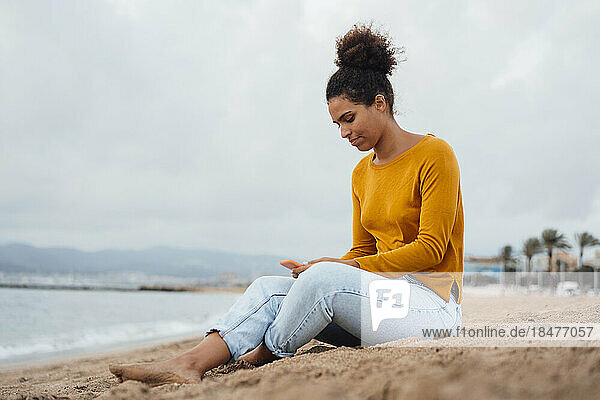 Junge Frau benutzt Mobiltelefon und sitzt auf Sand