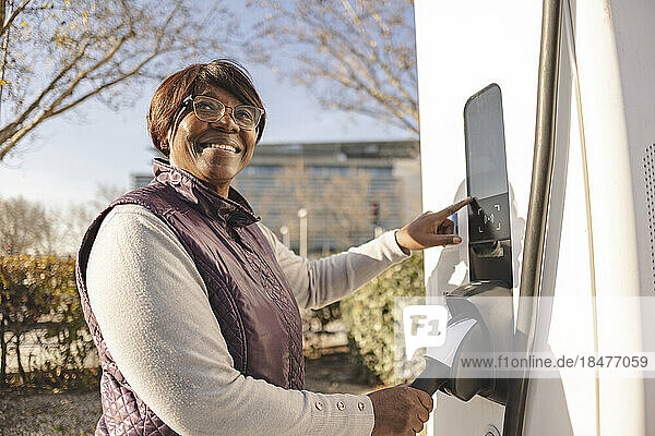 Lächelnde  nachdenkliche ältere Frau  die an der Elektroladestation steht