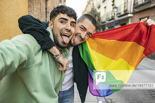 Happy gay couple sticking out tongues with rainbow flag on footpath