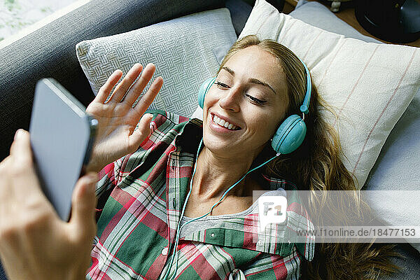Happy woman waving through smart phone wearing headphones lying on couch at home