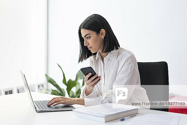 Young businesswoman with smart phone using laptop at desk