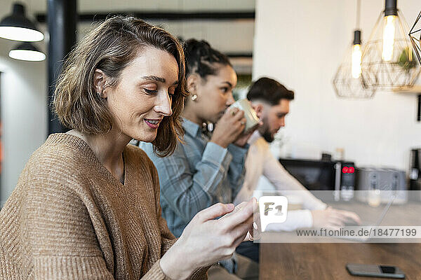 Lächelnde Geschäftsfrau benutzt Mobiltelefon mit Kollegen im Hintergrund