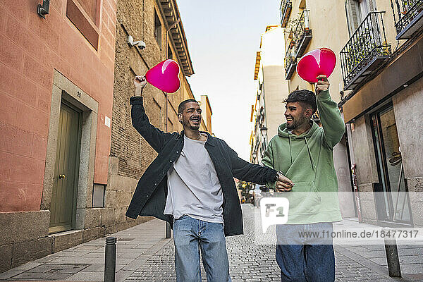 Happy gay couple having fun with red heart shape balloons amidst buildings at street