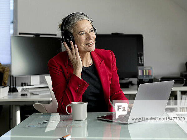 Happy senior businesswoman wearing wireless headphones sitting with laptop at desk