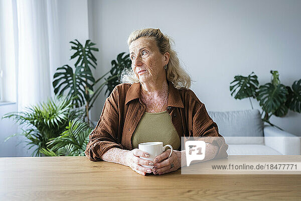 Sad elderly woman with mug sitting at home