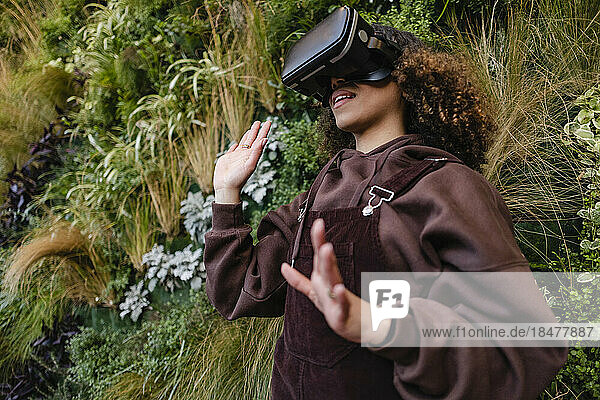Young woman gesturing with VR goggles in front of plants