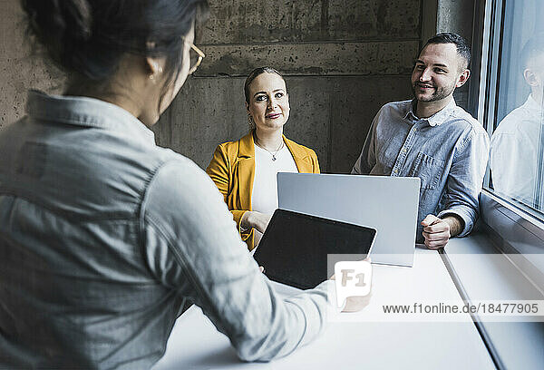 Lächelnde Kollegen schauen einer Geschäftsfrau zu  die bei einer Besprechung im Büro einen Tablet-PC benutzt