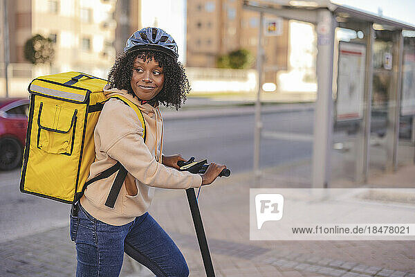 Smiling delivery woman with electric push scooter at footpath