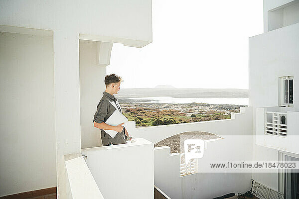 Nachdenklicher junger Mann steht mit Laptop auf dem Balkon