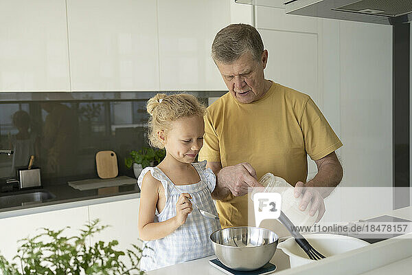 Senior man measuring flour by granddaughter in kitchen at home