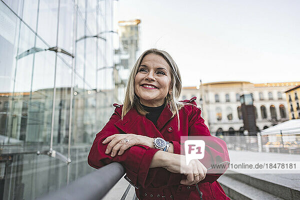 Smiling thoughtful mature woman leaning on railing outside modern building