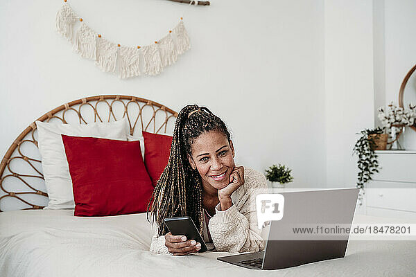 Smiling freelancer lying on bed with laptop and smart phone at home