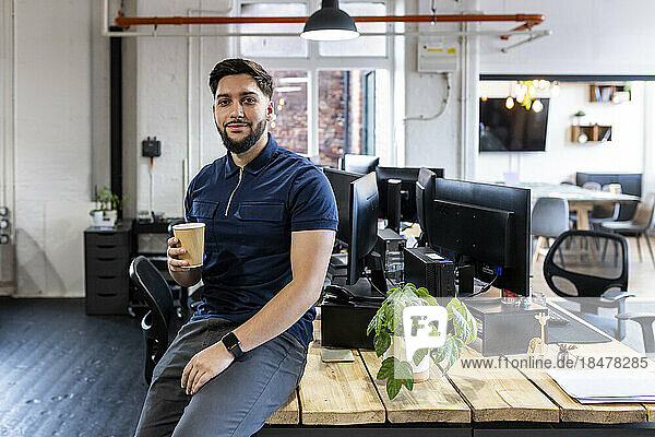 Smiling businessman with disposable cup sitting on desk at office