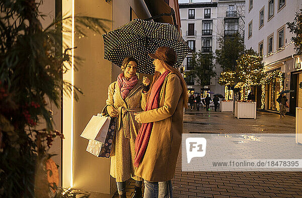 Smiling young woman holding umbrella and standing with friend on footpath