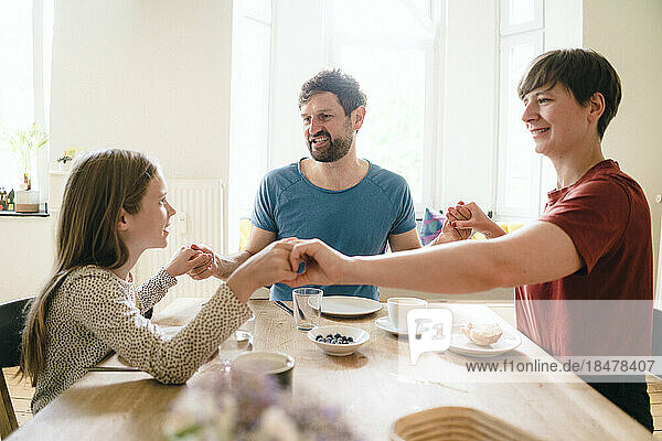 Father and mother holding hands with daughter sitting at dining table in home