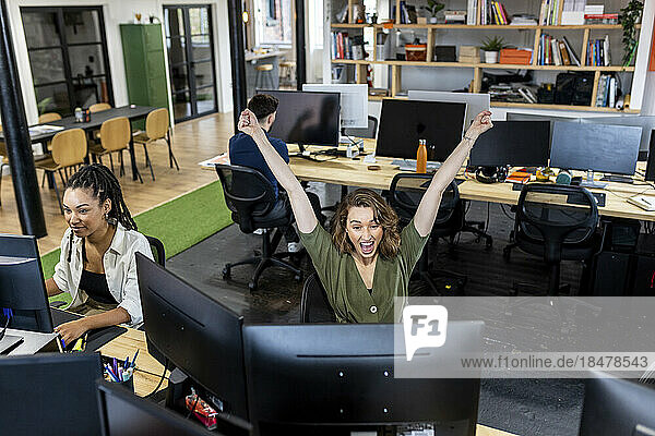 Happy businesswoman with arms raised working with colleagues in office