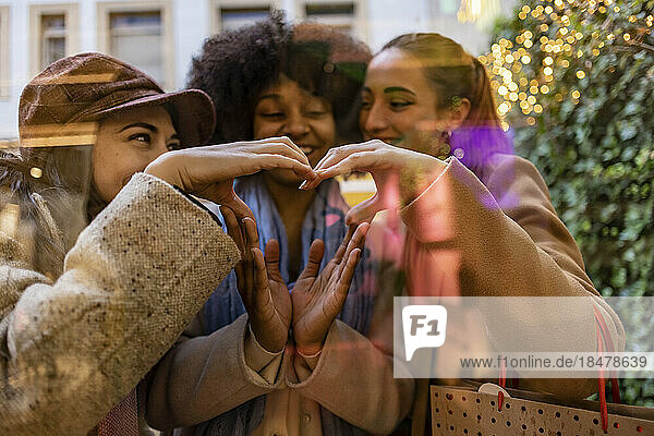 Happy friends making heart shape together with hands seen through glass