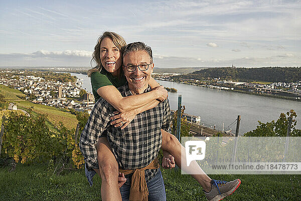 Happy man piggybacking woman by river under sky