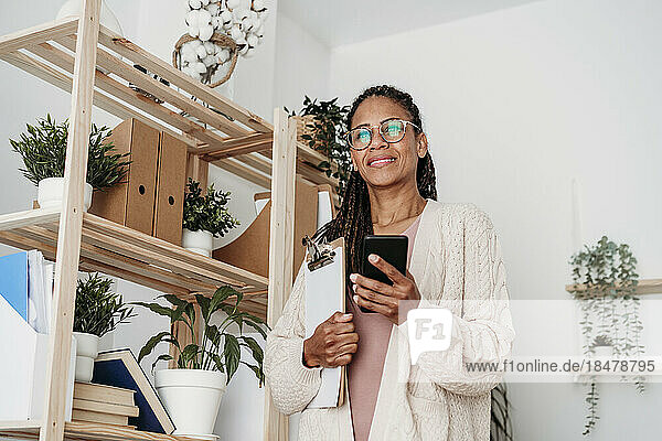 Smiling freelancer standing with clipboard and smart phone by shelf at home office