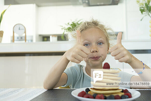 Girl making thumbs up gesture by plate of fresh pancakes at home