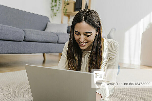 Glückliche junge Frau mit Laptop  die zu Hause auf dem Teppich liegt
