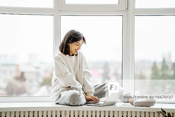 Happy woman using laptop sitting on window sill