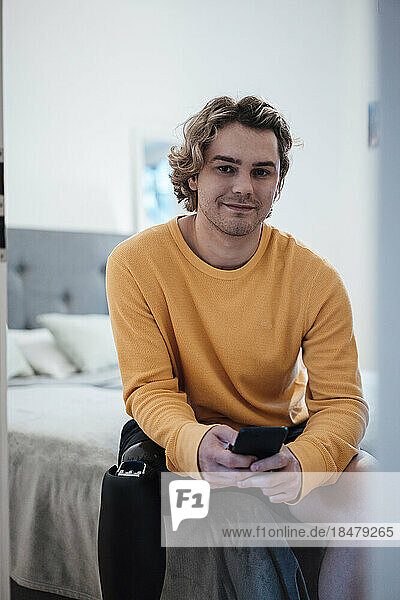 Lächelnder junger Mann mit Mobiltelefon sitzt zu Hause im Bett