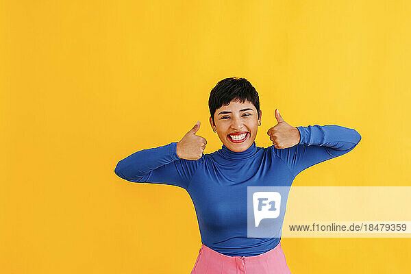 Happy young woman showing thumbs up against yellow background