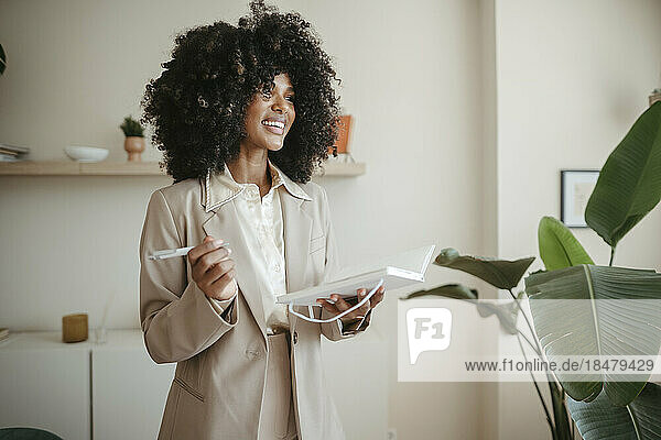 Happy businesswoman with Afro hairstyle standing with book in office