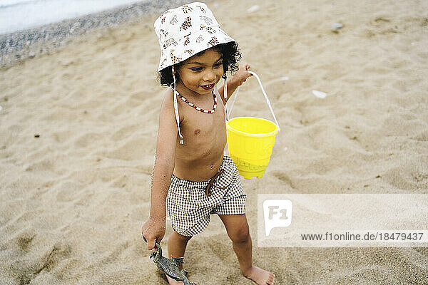 Cute boy wearing hat with bucket and toy on sand at beach