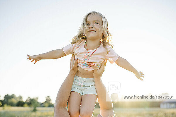 Mother lifting happy cute girl with arms outstretched in field on sunny day