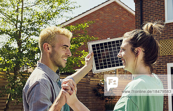 Smiling couple with hands intertwined holding solar panel in front of house