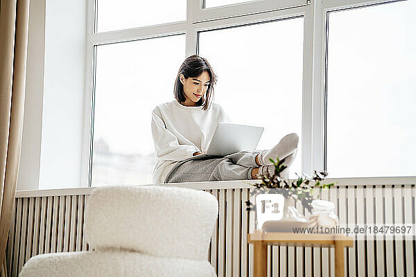 Lächelnde Frau mit Laptop  die zu Hause auf der Fensterbank sitzt