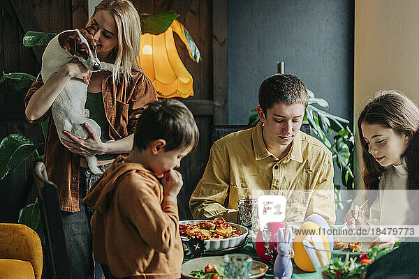 Happy woman holding dog with family having Easter dinner at home