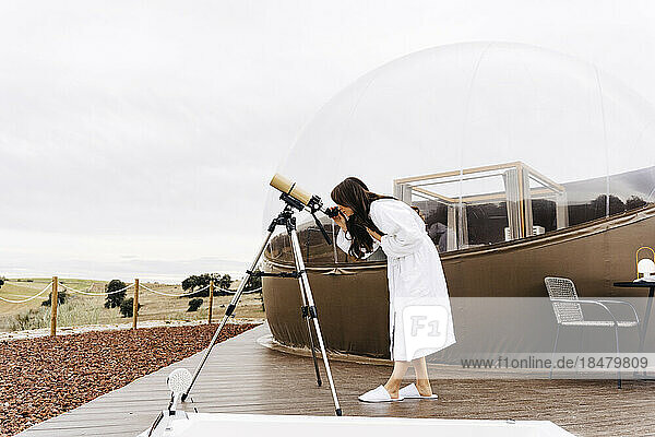 Young woman wearing bathrobe looking through telescope by dome tent