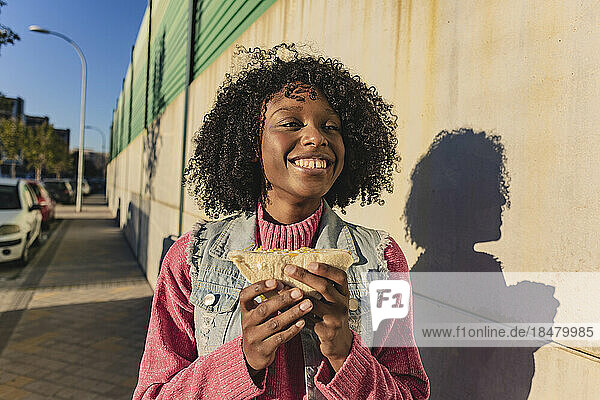 Happy woman holding sandwich standing at footpath