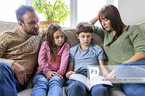 Boy reading book with family on sofa in living room