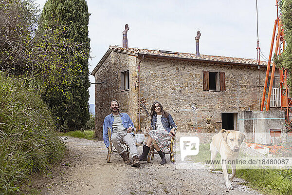 Happy mature couple sitting on chair with dog in front of house