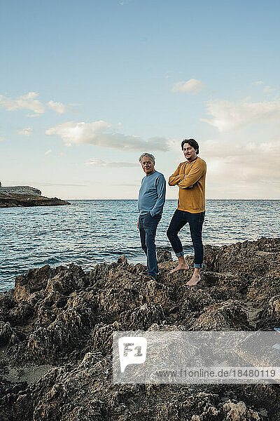 Senior man standing on rock with son in front of sea