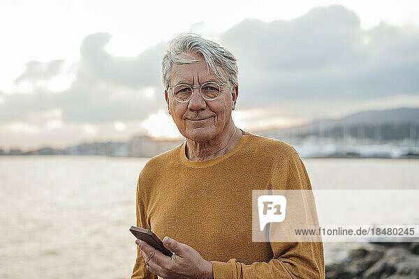 Smiling senior man standing with mobile phone in front of sea