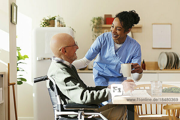 Smiling caretaker giving mug to senior man on wheelchair at home