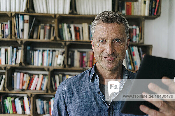 Lächelnder Mann mit Tablet-PC steht vor Bücherregal