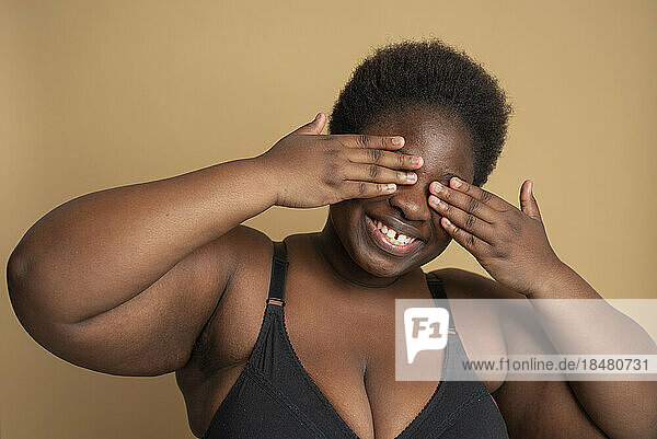 Smiling plus size woman in black bra covering her face with hands against beige background