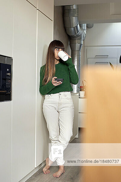 Frau trinkt Kaffee und steht mit Smartphone in der heimischen Küche