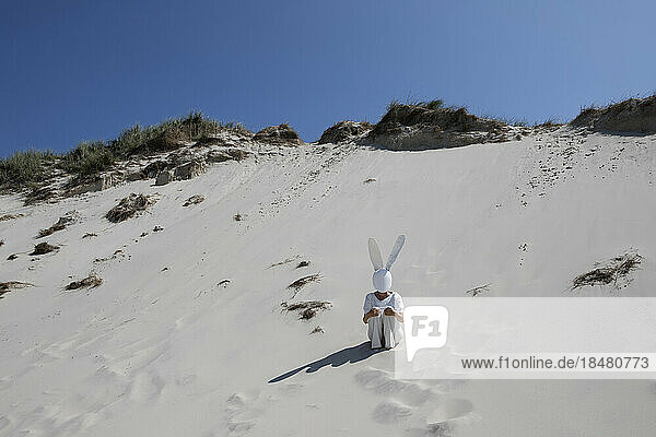 Frau mit Kaninchenmaske sitzt an sonnigem Tag auf Sanddüne