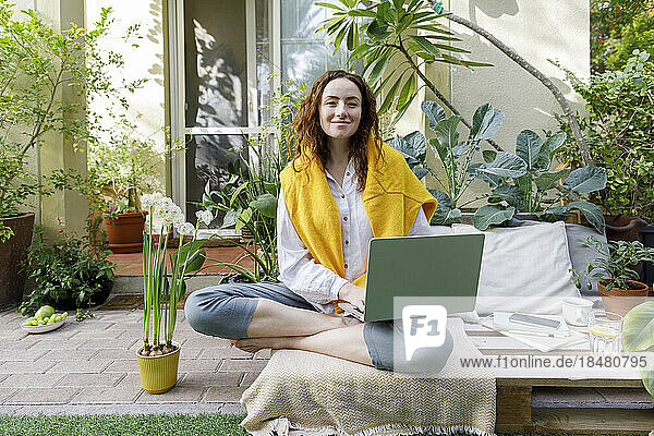 Freelancer sitting with laptop on wooden crate at backyard