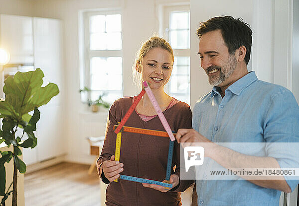 Smiling man talking to woman holding house model made of folding ruler at home