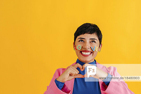 Cheerful young woman making heart shape with hands over yellow background