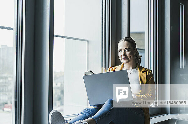 Lächelnde Geschäftsfrau mit Laptop sitzt am Fenster im Büro